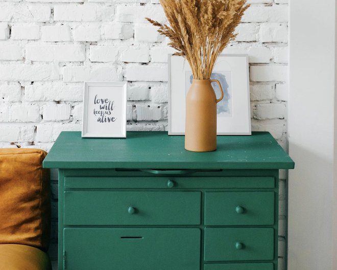 Framed pictures and a vase of wild grass on top of a chest of drawers against a whitewashed brick wall.
